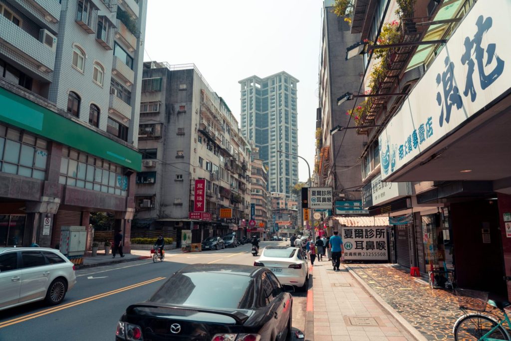 Lively city street with various buildings, cars, and pedestrians on a sunny day.